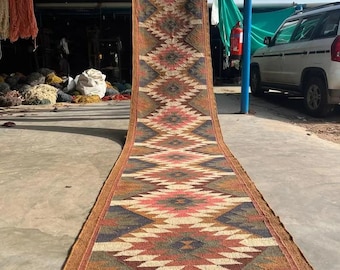 May include: A long, rectangular rug with a geometric pattern in shades of red, pink, brown, and blue. The rug has a brown border and a repeating diamond design. The rug is displayed outdoors.