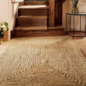 May include: A rectangular jute rug with a geometric design lies on a wooden floor. The rug is a natural beige color and has a textured, woven appearance. A staircase with brown carpet and wooden steps is in the background.