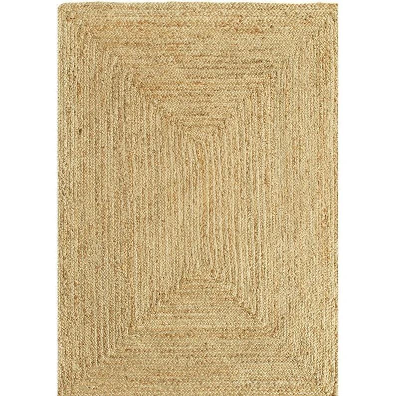 May include: Rectangular jute rug with a natural, light brown color. The rug features a woven design with a rectangular border and a central square pattern. The rug is suitable for indoor use.