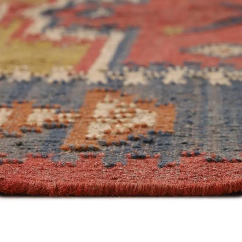 May include: Close-up of a woven rug with a geometric pattern. The rug features a color palette of red, blue, beige, and yellow. The texture of the rug is visible, with a slightly raised pile. The rug's edge is a solid red.