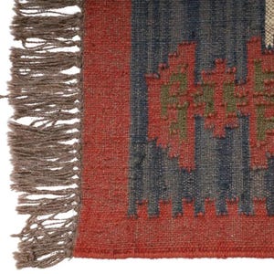 May include: Close-up of a woven rug with a geometric pattern. The rug features a red border and accents, with a central design in blue and green. The rug has a fringed edge in a neutral brown color. The rug is made of a textured material.