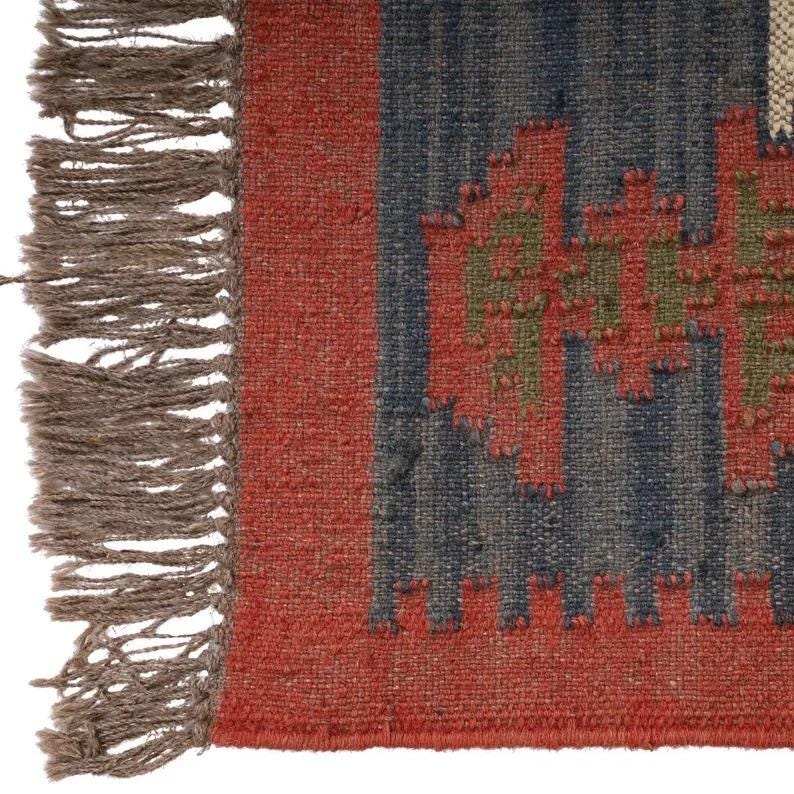 May include: Close-up of a woven rug with a geometric pattern. The rug features a red border and accents, with a central design in blue and green. The rug has a fringed edge in a neutral brown color. The rug is made of a textured material.