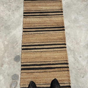 Può includere: Un tappeto rettangolare in juta con strisce nere orizzontali. Il tappeto è di colore marrone naturale e sembra essere tessuto a mano. Il tappeto è posizionato su una superficie di cemento. Le scarpe nere sono visibili in basso.