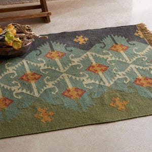 May include: A rectangular rug with a geometric pattern in shades of green, blue, and orange. The rug has a fringed edge and a repeating design of diamond and floral shapes. The rug is made of a woven material.