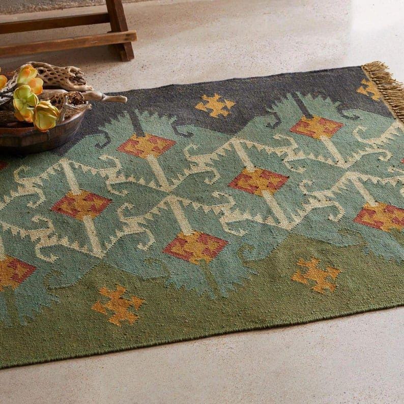 May include: A rectangular rug with a geometric pattern in shades of green, blue, and orange. The rug has a fringed edge and a repeating design of diamond and floral shapes. The rug is made of a woven material.