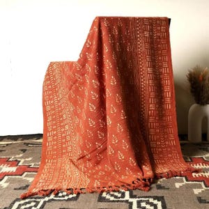 Puede incluir: Manta de color naranja quemado con patrones de hojas y geométricos blancos. La manta está drapeada sobre una silla, mostrando sus bordes con flecos y su tejido texturizado. La manta está hecha de un material suave.