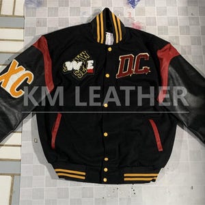 Puede incluir: Chaqueta universitaria negra con mangas de cuero negro, con detalles en amarillo y rojo. La chaqueta tiene el texto "XC" en la manga izquierda y "DC" en el pecho. La chaqueta también tiene el texto "WWE SE" en el pecho. Cierre de botones.
