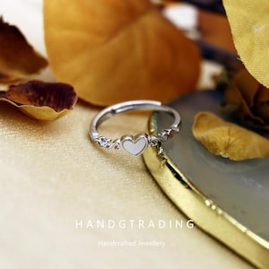 May include: A silver ring featuring a white heart-shaped centre. The ring has a delicate design with small decorative elements on the band. The image also shows the text "HANDGTRADING" and "Handcrafted Jewellery".