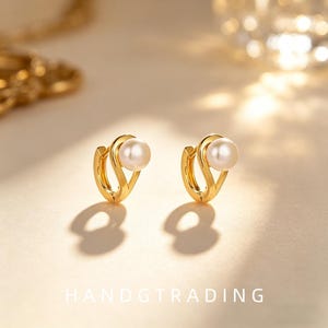 May include: A pair of gold hoop earrings featuring a smooth, curved metal design with a single round white pearl accent on each piece. The text HANDGTRADING is visible at the bottom of the frame, centered below the two earrings.