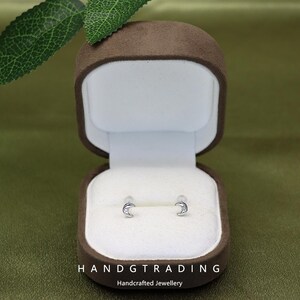 May include: Silver crescent moon stud earrings with a small gemstone, presented in an open brown jewellery box with a white interior. The box is set against a green backdrop. The text "HANDGTRADING Handcrafted Jewellery" is at the bottom.