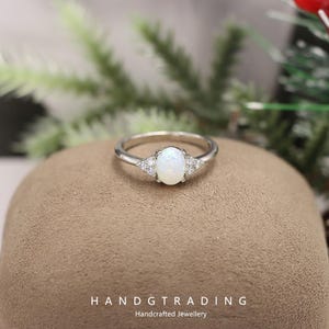 Könnte beinhalten: Ein silberner Ring mit einem ovalen, weißen Opal-Mittelstein und kleinen, dreieckigen Clustern aus klaren Steinen auf jeder Seite. Der Ring liegt auf einer braunen Oberfläche. Der Text lautet "HANDGTRADING Handcrafted Jewellery".