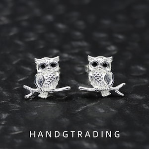 May include: A pair of silver-toned owl stud earrings, each featuring a detailed owl perched on a branch with small black circular eyes. The text HANDGTRADING is printed in white at the bottom of the image, centred below the two owl earrings.