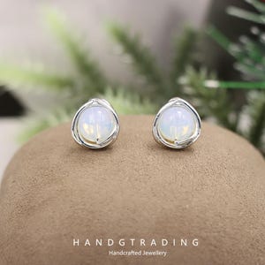 May include: Two stud earrings with a round, opalescent stone set in a silver-toned, twisted metal frame. The earrings are presented on a brown surface. Text at the bottom reads "HANDGTRADING Handcrafted Jewellery".