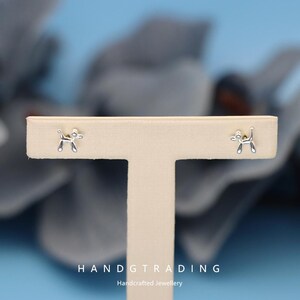 May include: A pair of silver-colored stud earrings shaped like balloon dogs. The earrings are displayed on a beige jewelry stand against a blurred blue background. The text "HANDTRADING Handcrafted Jewellery" is visible at the bottom.