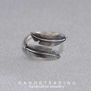 May include: A silver ring with a feather design. The ring has two detailed feather accents. The ring is set against a light grey background. The text "HANDGTRADING Handcrafted Jewellery" is at the bottom. The ring is a piece of handcrafted jewellery.