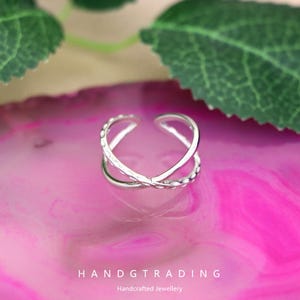 Könnte beinhalten: Verstellbarer Ring in Silber mit einem X-förmigen Design. Der Ring hat ein strukturiertes Muster auf dem Band und wird auf einer rosa Oberfläche mit grünen Blättern im Hintergrund gezeigt. Der Text "HANDGTRADING" und "Handcrafted Jewellery" befindet sich unten.