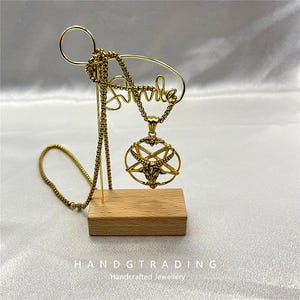 Puede incluir: Un collar dorado con un colgante con un diseño de estrella de cinco puntas y cabeza de cabra. El collar se exhibe en un soporte dorado con la palabra "smile" en cursiva. El soporte está sobre una base de madera. El texto "HANDTRADING Handcrafted Jewellery" está en la parte inferior.