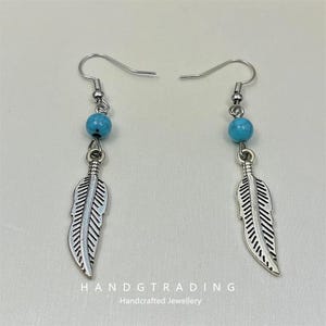 Puede incluir: Un par de pendientes de pluma plateados con cuentas de color turquesa. Los pendientes tienen un cierre de gancho. El texto "HANDGTRADING Handcrafted Jewellery" se encuentra en la parte inferior de la imagen.