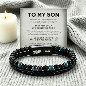Peut inclure: Un bracelet noir à perles avec des accents bleus et marron, avec un bracelet en cuir tressé. Le bracelet a un fermoir magnétique avec le texte "SHIELDED BY LOVE" et "J.B.♥". Une carte avec le texte "TO MY SON" est en arrière-plan.