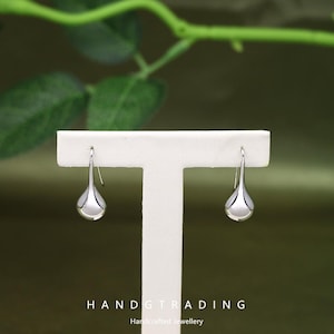 May include: A pair of silver teardrop earrings. The earrings are displayed on a white stand against a blurred green background. The earrings have a polished, reflective surface. The text "HANDGTRADING Handcrafted Jewellery" is at the bottom.