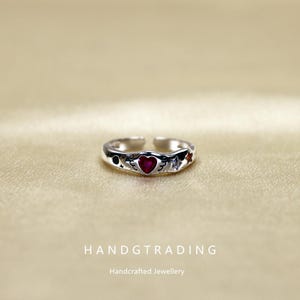 May include: A silver-toned ring with a heart-shaped red gemstone at the center. The ring features celestial and wing designs on the band. The text "HANDGTRADING" and "Handcrafted Jewellery" are at the bottom.