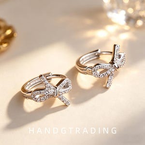 May include: A pair of silver-toned hoop earrings featuring a bow-shaped charm encrusted with small clear stones. The earrings have a hinged closure mechanism and are positioned on a light surface with the text HANDGTRADING visible at the bottom.