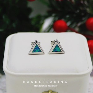 May include: Two silver triangle stud earrings with a blue-green inlay. The earrings are presented on a white surface. The text "HANDGTRADING Handcrafted Jewellery" is below. The background has blurred green foliage and red berries.