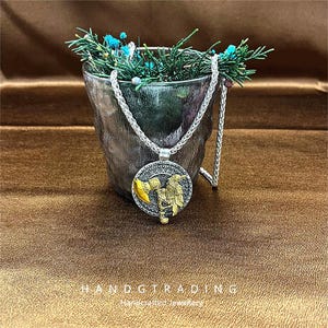 May include: A silver-toned necklace with a circular pendant featuring a detailed design with gold and silver accents. The pendant depicts a figure holding a hammer and a flag. The necklace is displayed in front of a small, textured bucket with greenery.