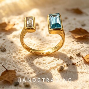 May include: A gold-toned open ring featuring two rectangular stones, one clear and one light blue, set in a bezel style. The band has an engraved texture and the text HANDGTRADING is visible at the bottom of the frame in white capital letters.