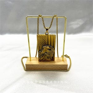 Könnte beinhalten: Eine goldfarbene Halskette mit Anhänger, die eine amerikanische Flagge und einen Adler zeigt. Der Anhänger hängt an einer Kette und wird auf einem Holzsockel mit einem goldenen Ständer präsentiert. Die Halskette ist detailliert gearbeitet.