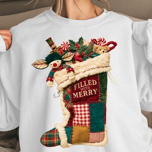 May include: A white long-sleeve sweatshirt featuring a graphic of a patchwork Christmas stocking filled with toys, including a nutcracker and teddy bear. The stocking has a cream-colored faux fur cuff and the text FILLED WITH MERRY.