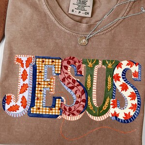 May include: A brown short-sleeved t-shirt with a crew neckline and a Comfort Colors label. The front features the word JESUS in large block letters, each filled with distinct autumn-themed patterns including orange leaves, acorns, and wheat.