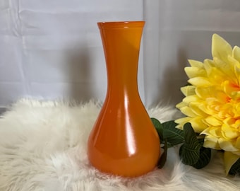 May include: An orange glass vase with a flared top and a rounded base. The vase is a vibrant shade of orange and is set against a white background. A yellow flower is visible in the background.
