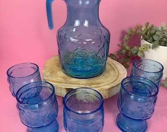 May include: A vintage blue glass pitcher with a handle and lid, surrounded by six matching glasses. The glassware features a floral design and sits on a wooden surface against a pink backdrop. The set is ideal for serving beverages.