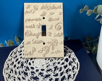 May include: A beige ceramic light switch cover with embossed floral and script designs. The cover is placed on a white crocheted doily. The background is a dark blue surface.