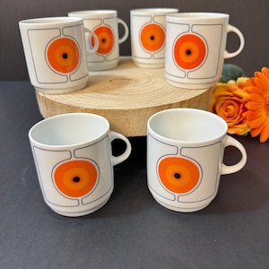 Op de afbeelding: Een set van zes witte keramische mokken met een retro-ontwerp. Elke mok heeft een grote oranje en zwarte cirkelvormige afbeelding in een zwart omkaderde vierkant. De mokken zijn gerangschikt op een houten oppervlak, met een donkere achtergrond en oranje bloemen.