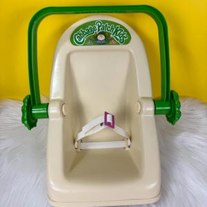 May include: A vintage Cabbage Patch Kids doll carrier in cream and green. The carrier has a green handle and accents, with the Cabbage Patch Kids logo on the front. It features a white strap with a pink buckle for securing a doll.
