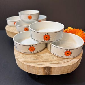 May include: A collection of six white ceramic bowls with a retro design. Each bowl has a black border and a central orange and yellow circular design. The bowls are arranged on a wooden slab, with an orange flower in the background.