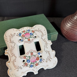 以下が含まれることがあります： 白い磁器製のダブルライトスイッチカバー、花柄のデザイン。 ピンク、青、赤の花、金色のトリム、装飾的なスクロールワークが特徴です。 装飾的な家の装飾アイテムです。