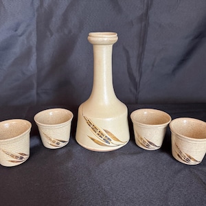 May include: A beige ceramic sake set featuring a tall, slender bottle and four small cups. The bottle and cups have a speckled texture and are decorated with a brown leaf design. The set is displayed on a dark surface.