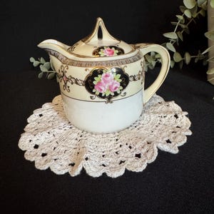 Könnte beinhalten: Eine cremefarbene Vintage-Sahnekännchen aus Porzellan mit Deckel, verziert mit einem schwarzen und goldenen Blumendesign mit rosa Rosen. Das Sahnekännchen steht auf einer weißen, gehäkelten Untersetzer. Griff und Ausguss sind sichtbar. Der Hintergrund ist schwarz.