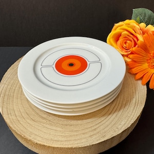 May include: Four white ceramic plates with a retro design, stacked on a wooden base. Each plate has a central orange and black bullseye design, framed by a black line. The plates are surrounded by orange and yellow flowers. The wooden base is approximately 20 cm in diameter.