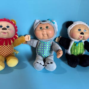 May include: Three Cabbage Patch Kids dolls dressed as animals. The lion doll is yellow and red with a red mane. The koala doll is grey with a blue bow. The panda doll is black and white with a green vest and blue bow.