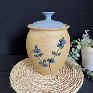 Peut inclure: Un pot en céramique beige avec un couvercle bleu clair et un bouton assorti. Le pot est décoré d'un motif de branche de myrtilles en bleu et vert. Le pot repose sur un dessous de plat tressé.