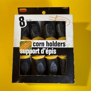 May include: A pack of eight corn holders. The holders are black with yellow tops, presented in a cardboard box. The box features the words "corn holders" and "support d'épis".