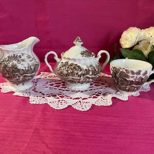 May include: A white porcelain tea set with a brown transferware design depicting a rural scene. The set includes a creamer, a lidded sugar bowl, and a teacup, all on a white lace doily. A bouquet of white and yellow roses is in the background.