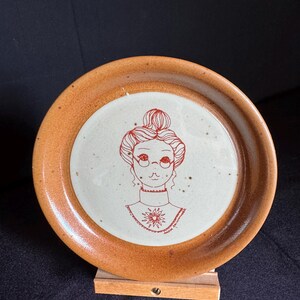 Peut inclure: Une assiette en céramique avec un bord marron et un centre blanc, ornée d'une illustration rouge d'une femme avec des lunettes et un chignon. L'assiette est présentée sur un petit support en bois, mettant en valeur un design unique et artisanal.