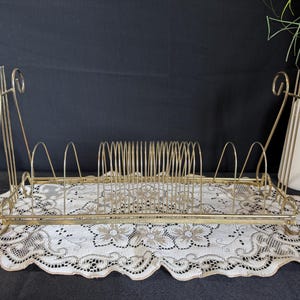 Puede incluir: Un portadiscos vintage de metal dorado con base rectangular y divisores verticales. El soporte tiene adornos decorativos y se apoya sobre un tapete de encaje blanco. Un jarrón blanco con vegetación se encuentra en el fondo.