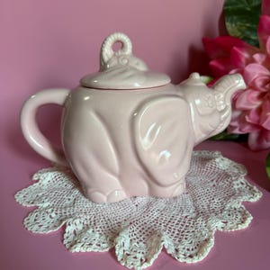 Peut inclure: Une théière en céramique rose clair en forme d'éléphant, avec un couvercle et une anse. Elle repose sur un dessous de plat en dentelle blanche festonnée. Le fond est rose, avec une fleur rose visible. Un objet décoratif charmant.