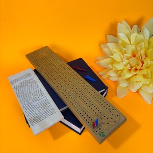 May include: A wooden cribbage board with holes for pegs, alongside a blue notebook, instructions, and a yellow flower against an orange backdrop. The board features red and blue pegs.